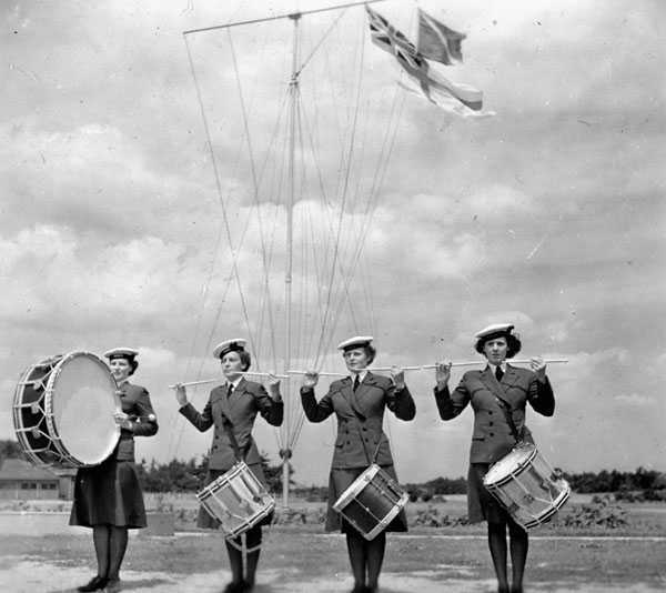Black and white photograph. 4 women in navy uniforms stand in a line. The furthest left plays a large base drum. The other three have snare drums, their sticks held horizontally in the air. The Red Ensign flies behind them.
